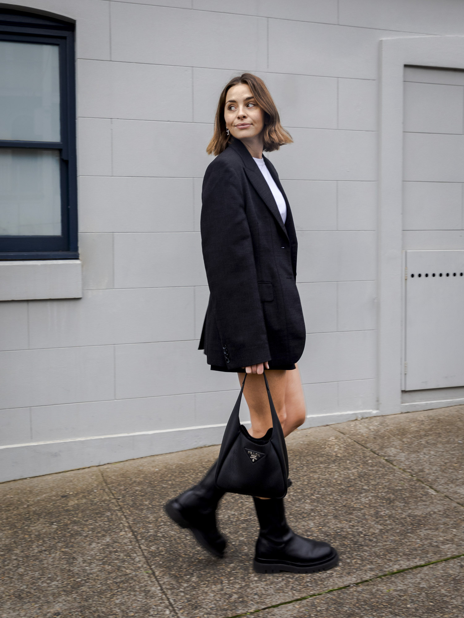 A woman in a black blazer, white shirt, and boots, carrying a Prada handbag, walks on a sidewalk with a light gray building behind her.