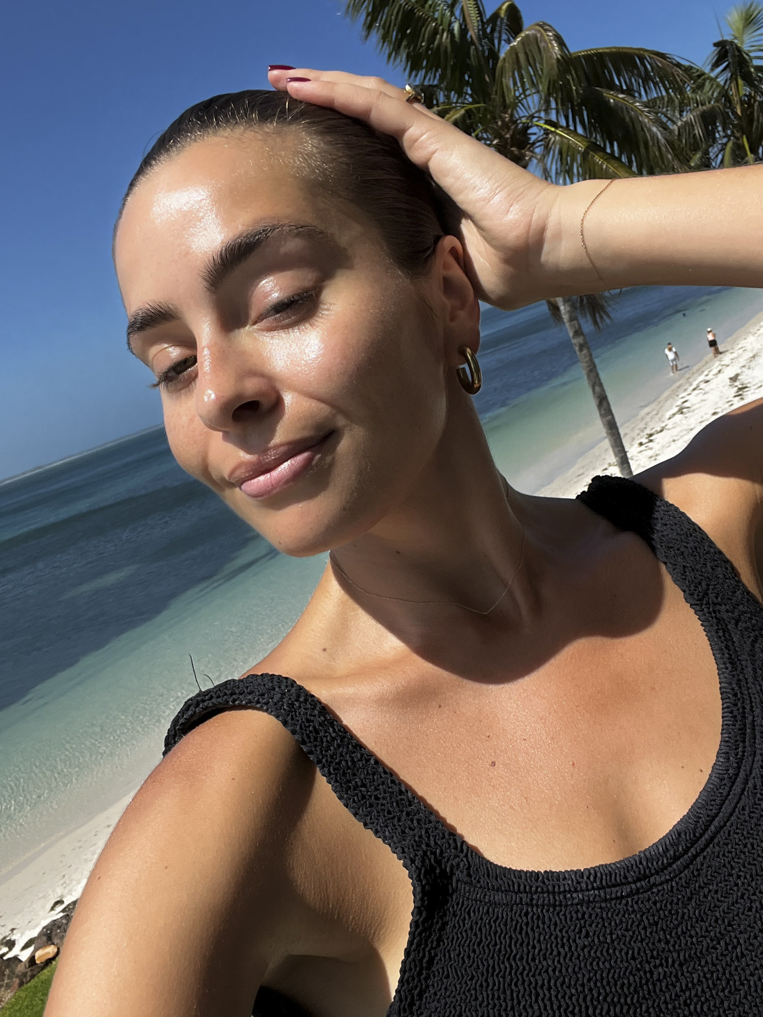 Adult female in black swimsuit poses on beach with palm trees and turquoise sea under sunny blue sky, enjoying a tropical vacation.