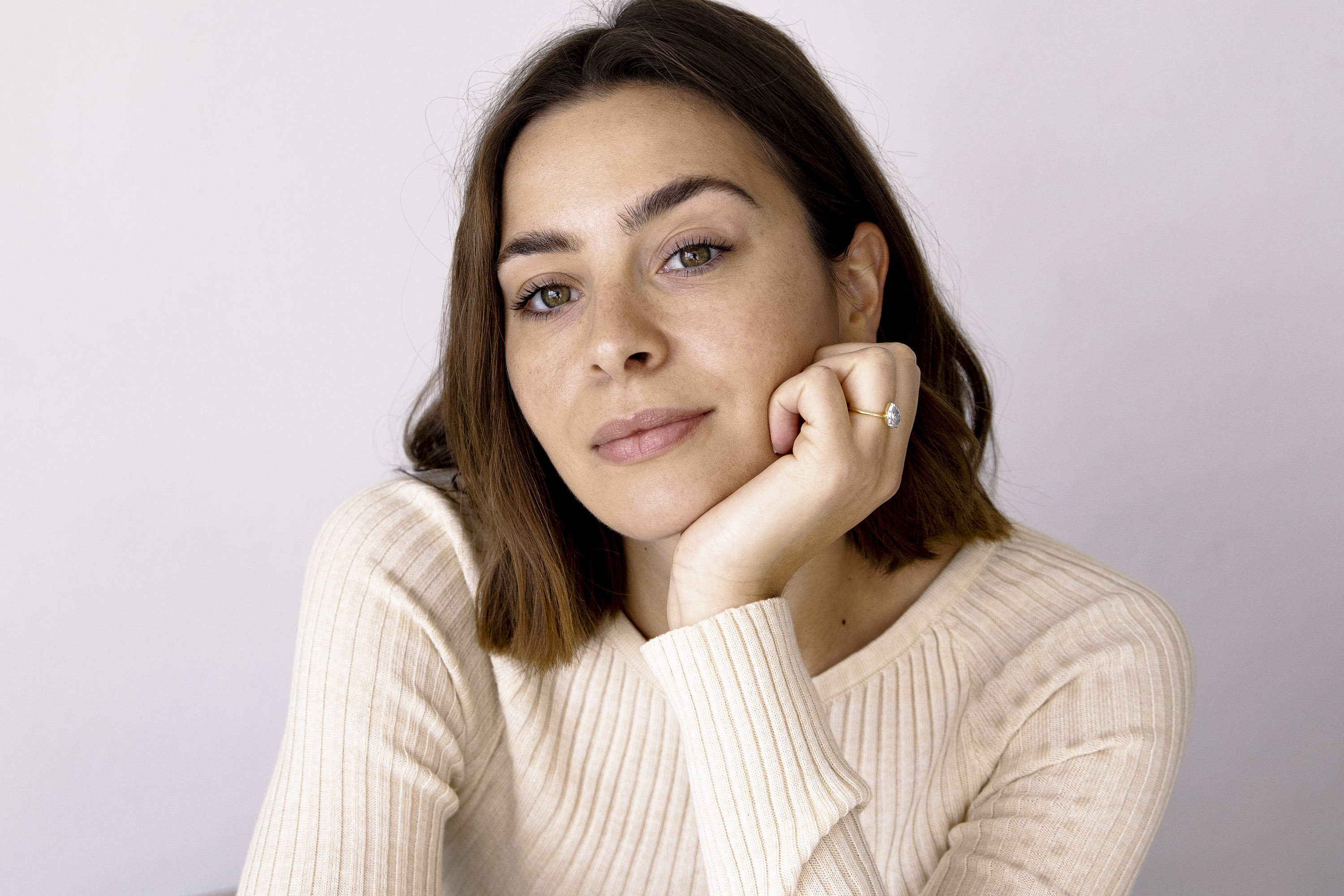 Close-up portrait of a woman with auburn hair wearing a cream-colored sweater, resting her chin on her hand against a plain white background.