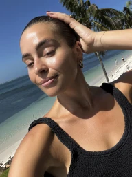 Close-up selfie of a woman in a black textured swimsuit smiling outdoors on a sunny tropical beach with palm trees and ocean.