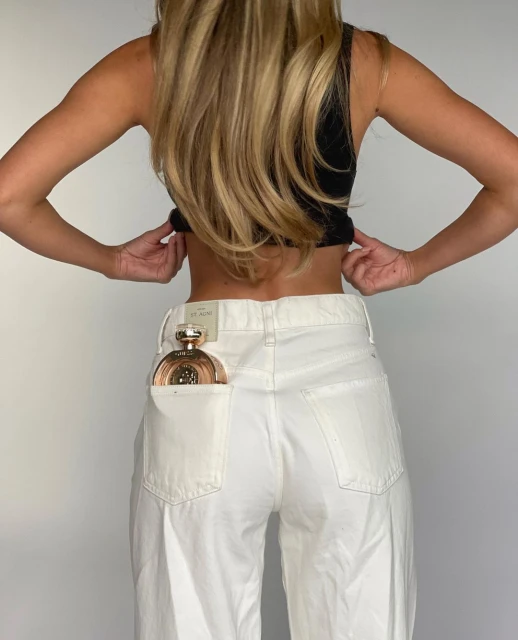 Blonde woman in black top and white jeans poses in a studio with a gold perfume bottle in her back pocket in a portrait shot.