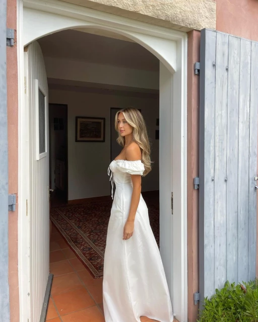A young blonde woman in a white off-shoulder dress stands in a doorway, looking away. Natural light illuminates the indoor scene.