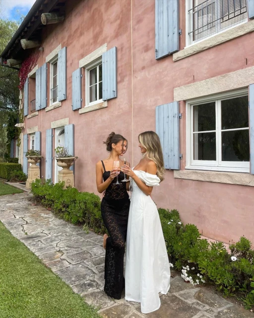 Two adult females in elegant dresses toasting with champagne in a garden setting next to a blush-colored building with blue shutters.