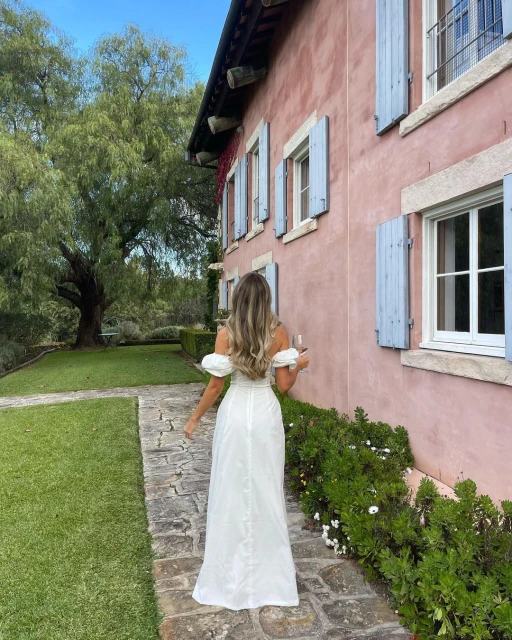 Back view of a woman in a white dress walking on a stone path beside a house with blue shutters and a green lawn and trees.