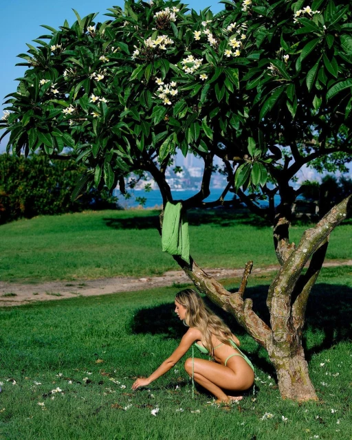 A woman in swimwear picks up white flowers that have fallen from a tree in a lush, green garden with a view of the ocean.