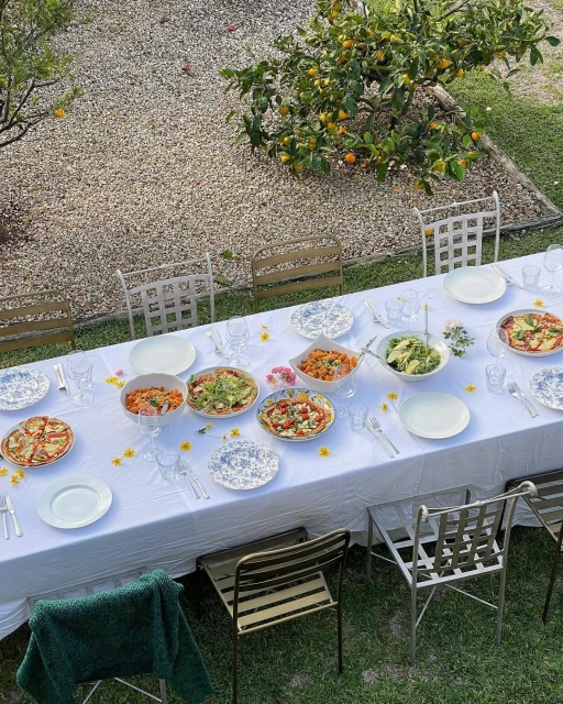 Outdoor dining table set with various bowls of food and plates, set under a tree with gravel ground covering in the background.