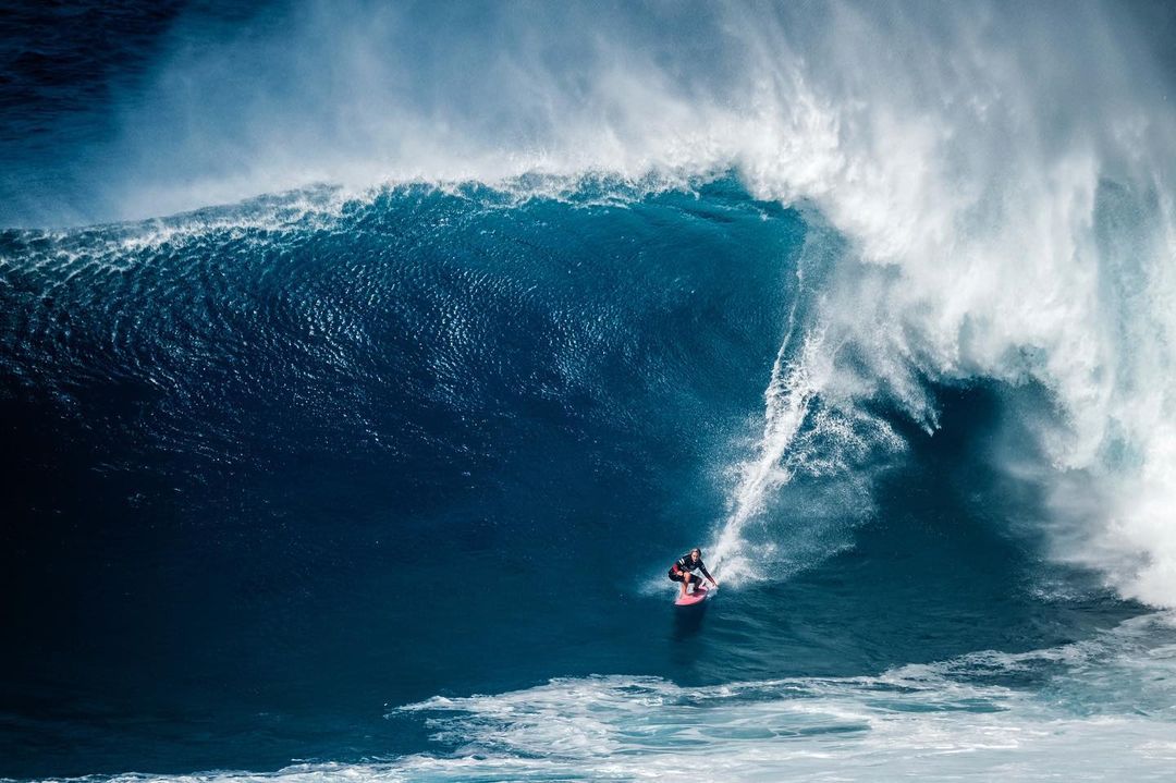 A surfer rides a massive blue ocean wave, with white water and foam creating a dramatic scene of power and skill in nature.