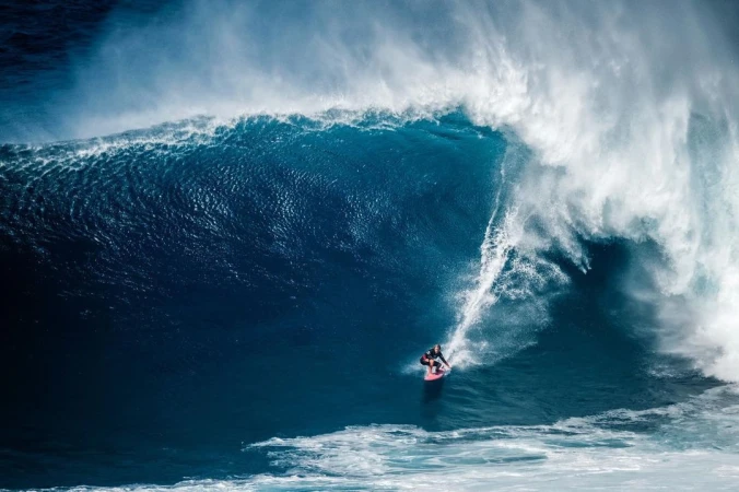 A surfer rides a massive blue ocean wave, with white water and foam creating a dramatic scene of power and skill in nature.