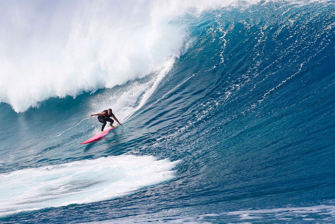 A surfer rides a pink surfboard down the face of a massive, blue ocean wave under bright, natural lighting conditions.