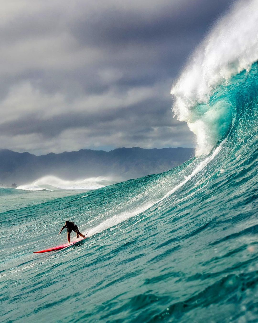 A surfer rides a large, turquoise ocean wave under a cloudy sky, showcasing the power of nature and a thrilling moment of athleticism.