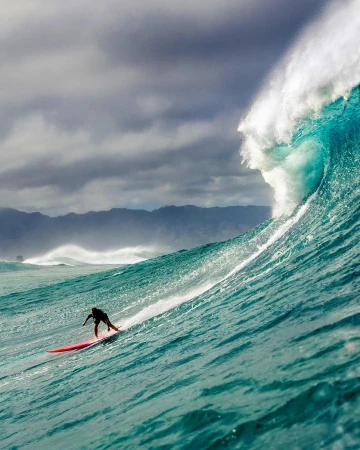 A surfer rides a large, turquoise ocean wave under a cloudy sky, showcasing the power of nature and a thrilling moment of athleticism.
