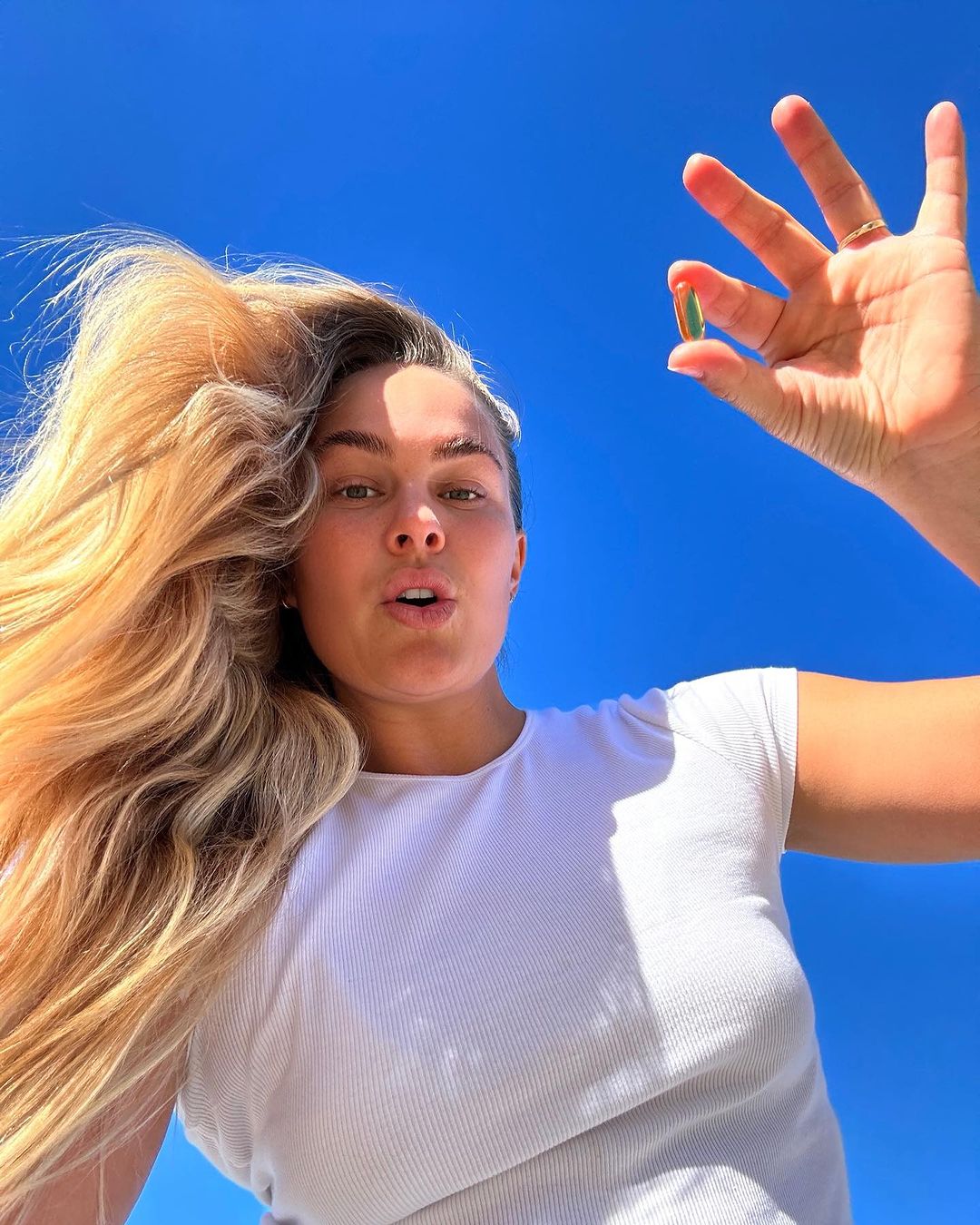 Woman holds an oblong capsule towards the camera, outdoors with a clear blue sky backdrop. Her long blonde hair and white shirt are visible.