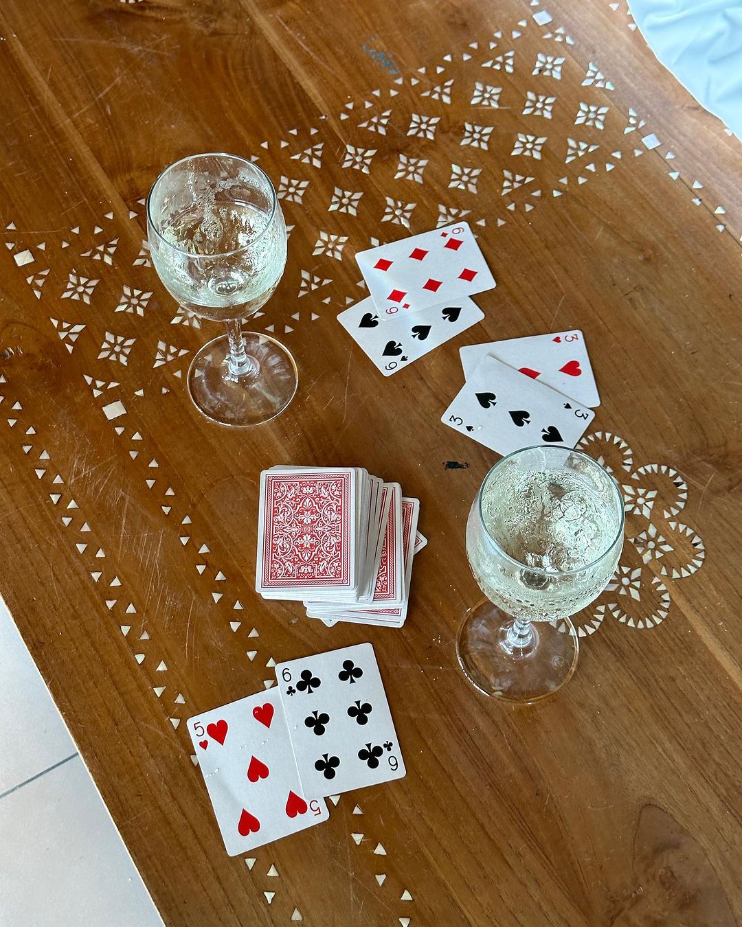 Overhead shot on wood table of two wine glasses, deck of cards, playing cards scattered, and patterned table inlay.