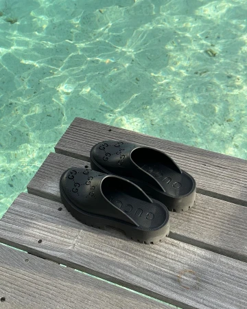 Black rubber platform slip-on clogs resting on a wooden dock above clear turquoise ocean water.
