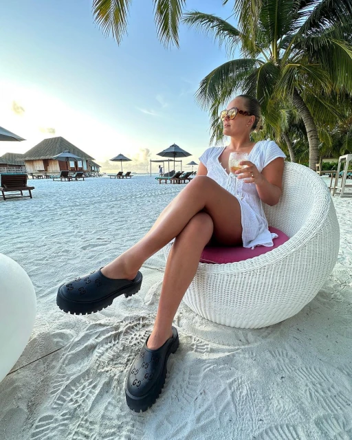 Woman in white dress sits in outdoor chair on white sand beach with drink and palm trees in background. Tropical vacation.