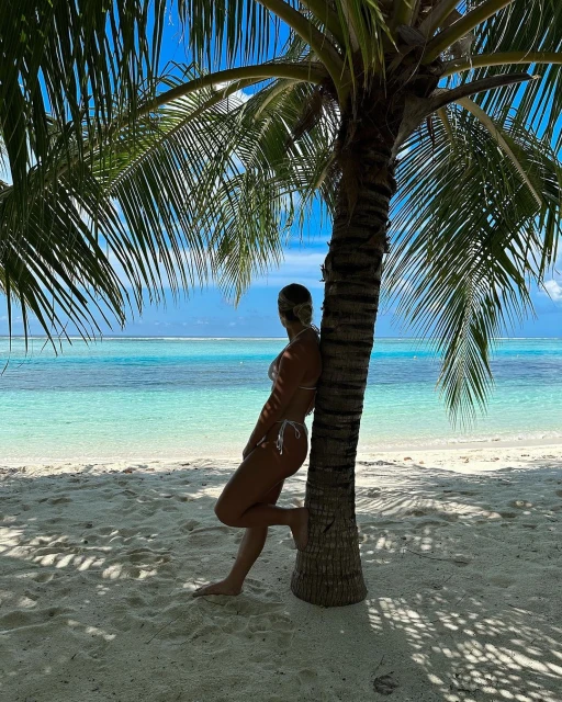 Woman in bikini poses under a palm tree on a white sand beach with turquoise ocean water in the background on a sunny day.