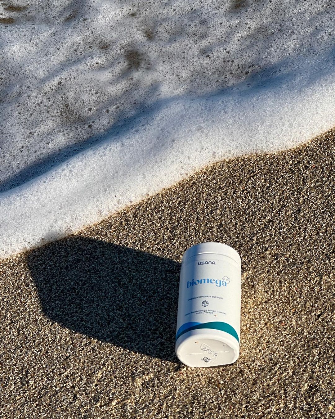 USANA Biomega fish oil supplement on a sandy beach with ocean foam in the background, product shot for health and wellness.
