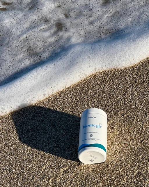 USANA Biomega fish oil supplement on a sandy beach with ocean foam in the background, product shot for health and wellness.