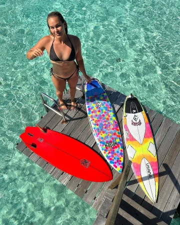 Woman in bikini stands on a wooden dock near three surfboards in turquoise water. Summer tropical scene for travel and watersports.