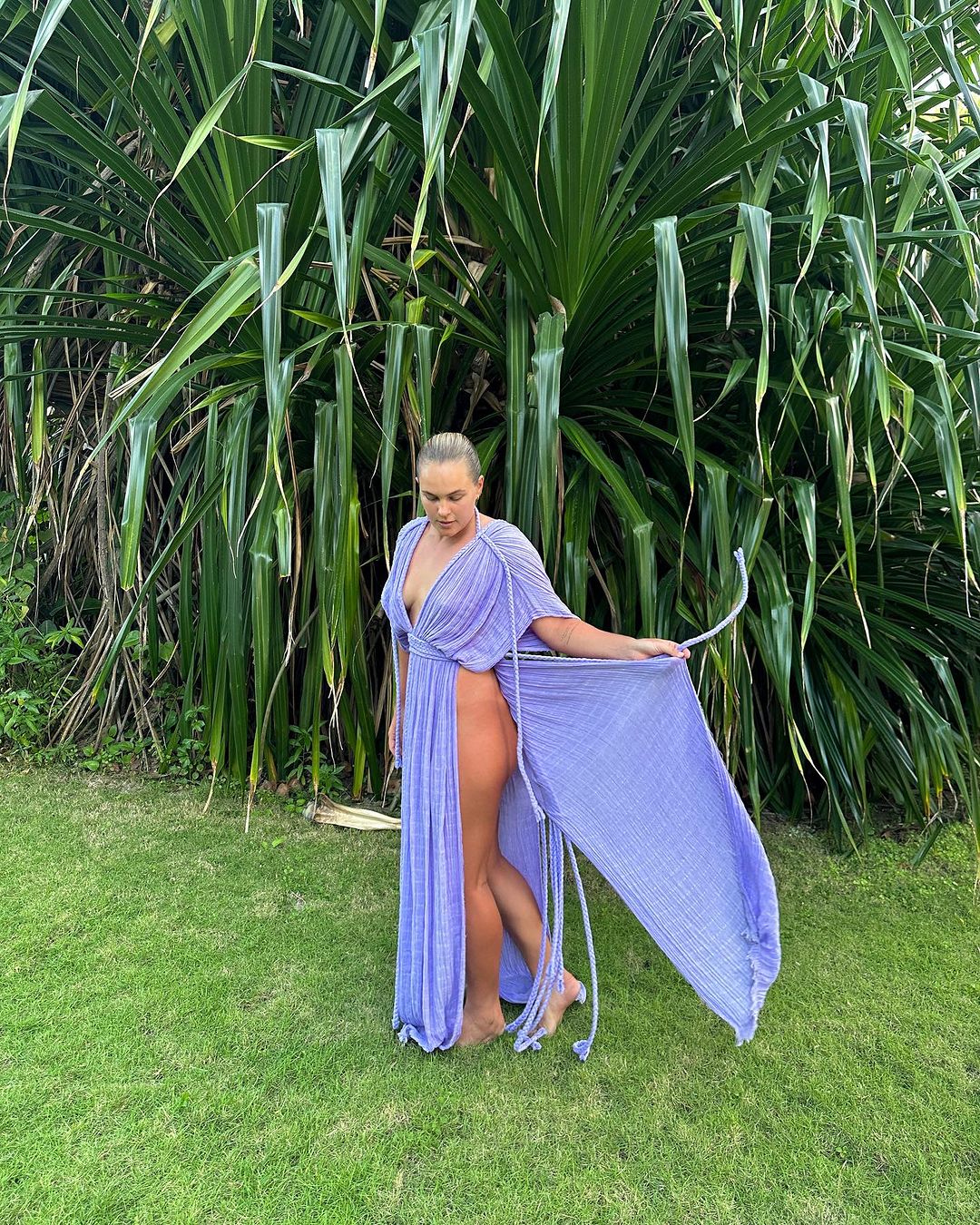 Woman poses in tropical location wearing periwinkle-blue dress with green tropical foliage, grass, and natural daylight background.