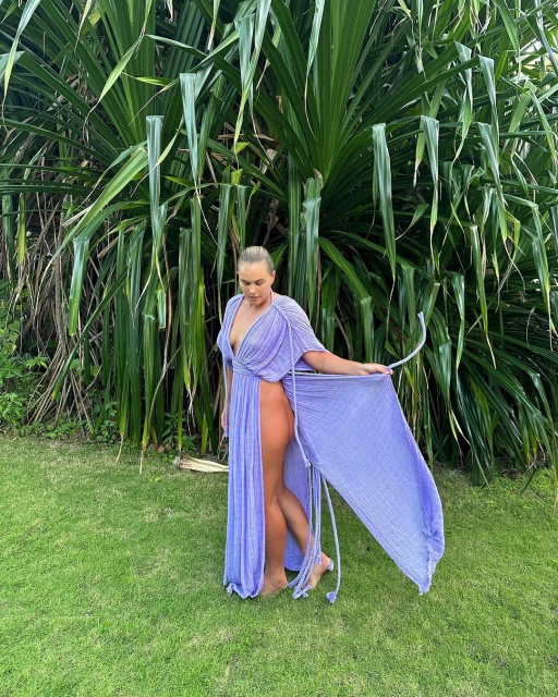 Woman poses in tropical location wearing periwinkle-blue dress with green tropical foliage, grass, and natural daylight background.