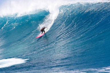 Surfer carving down a massive crashing blue ocean wave on a pink surfboard.