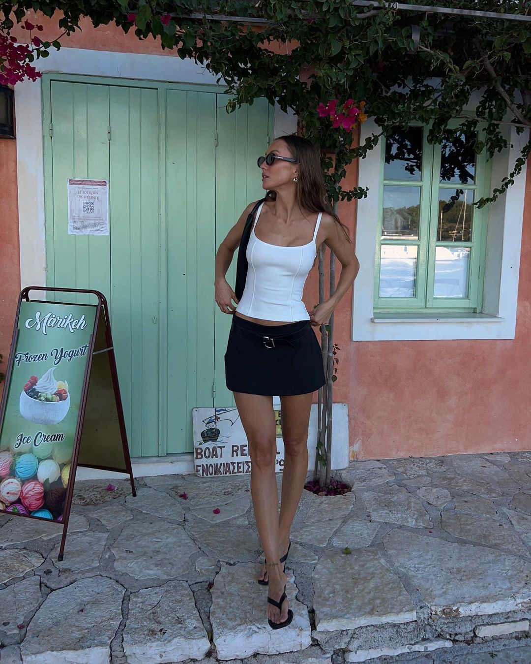 A young woman stands in front of a light green door and a pink wall, wearing a white tank top and black mini skirt, accessorized with sunglasses.