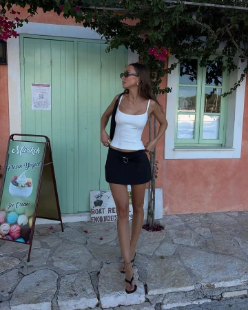 A young woman stands in front of a light green door and a pink wall, wearing a white tank top and black mini skirt, accessorized with sunglasses.