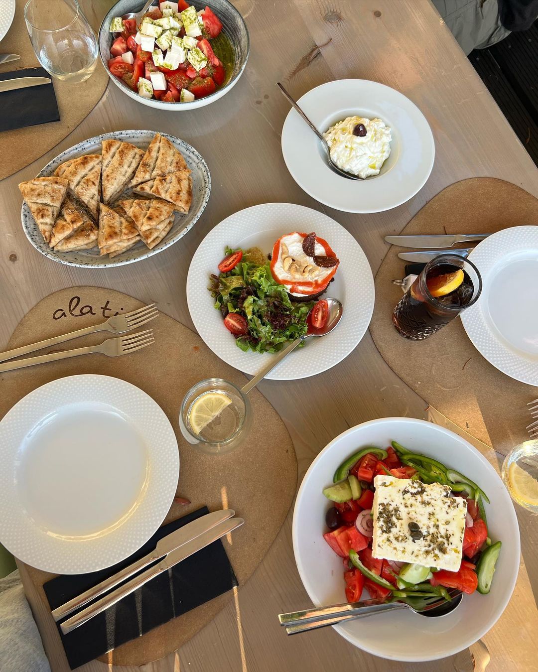 Overhead shot of a table with various plates of Greek salad, pita bread, and a beverage, showcasing a light and inviting dining experience.