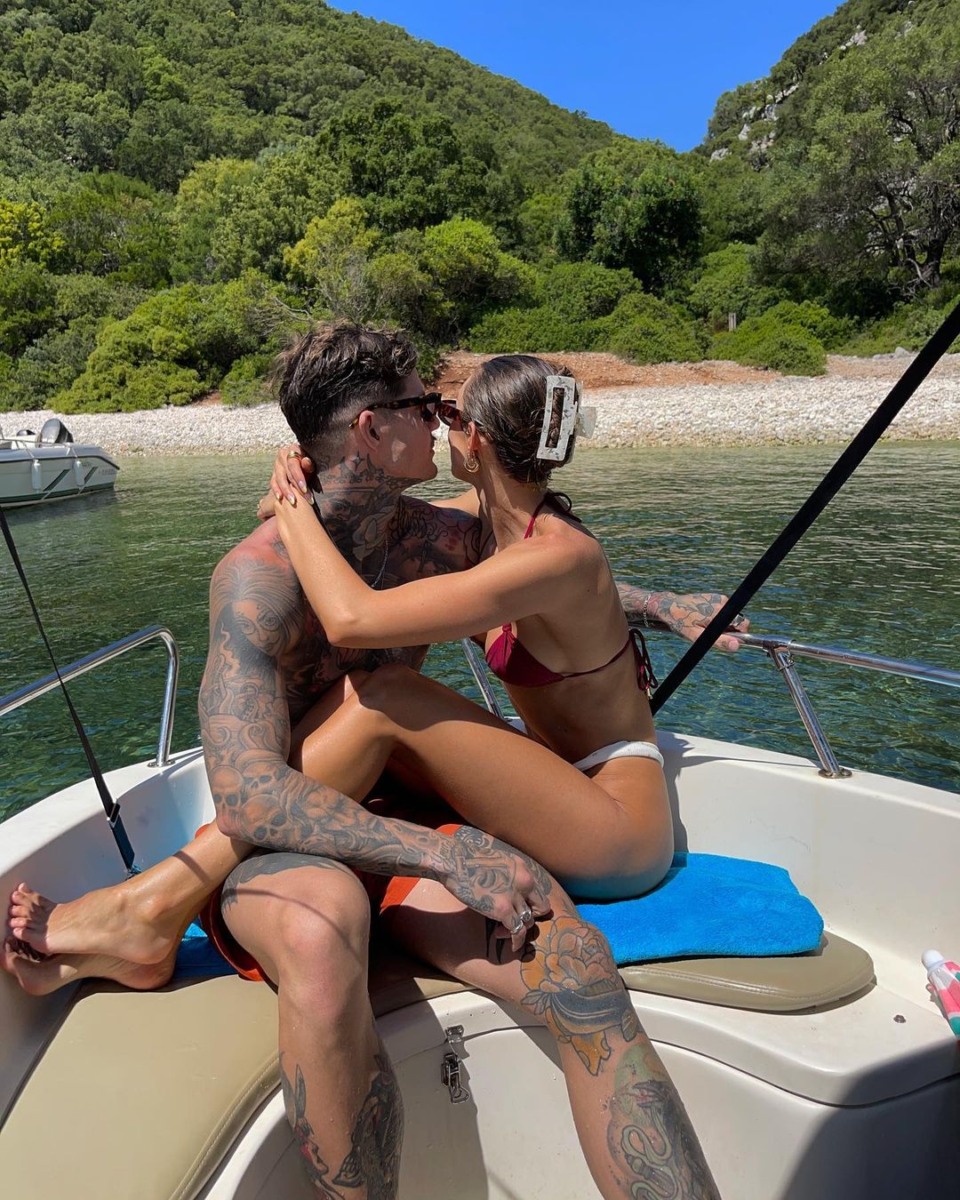 Couple embracing on a boat with forest backdrop. The woman wears a red bikini and the man is heavily tattooed. Water surrounds the boat.