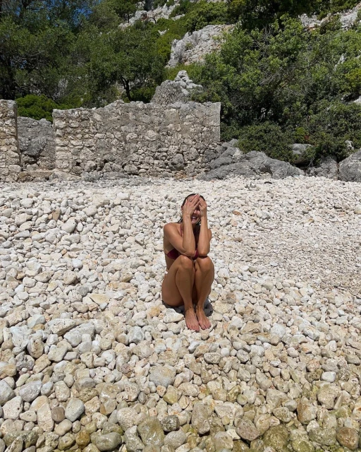 Woman in swimwear sitting on a pebble beach with her hands covering her face. Mountains and clear sky in the background.