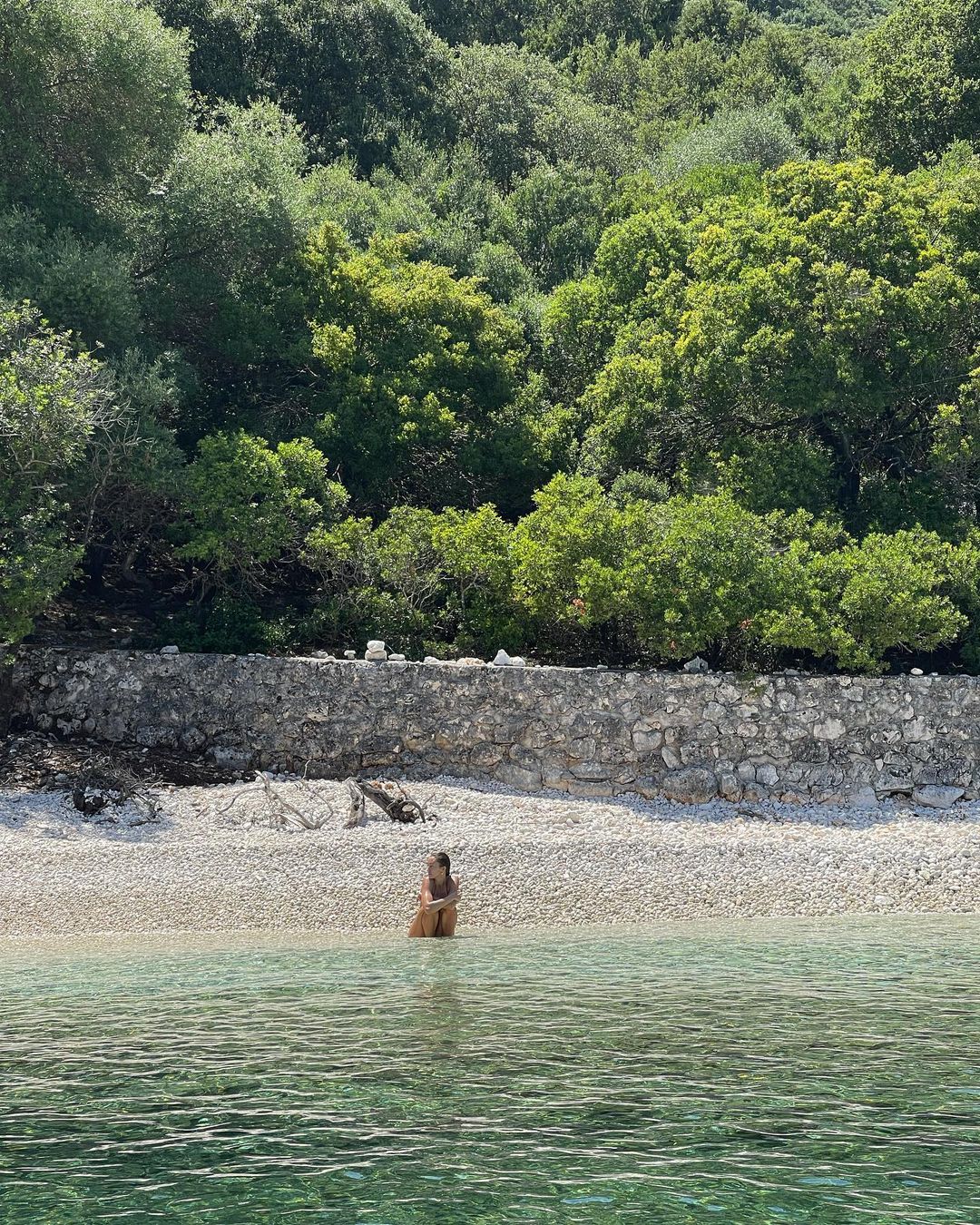 Woman seated in tranquil turquoise water at the shoreline of a pebbled beach backed by lush green forest under bright sunlight.