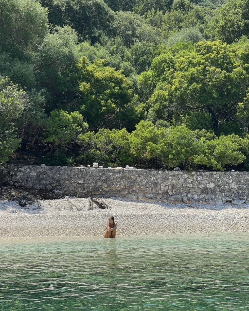 Woman seated in tranquil turquoise water at the shoreline of a pebbled beach backed by lush green forest under bright sunlight.