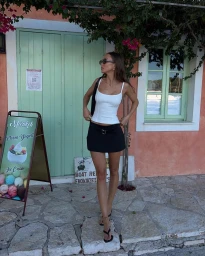Woman wearing a white tank top and black mini skirt posing outdoors in front of a peach-colored building with green doors and windows.