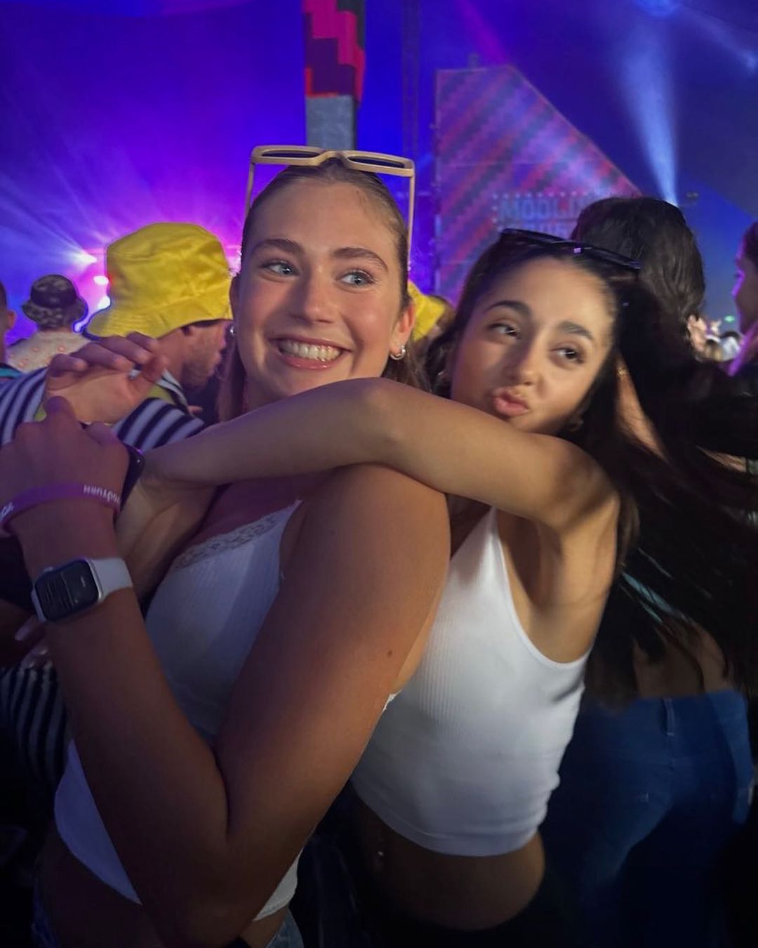 Two young adult women smile and hug at an outdoor music festival with crowd, stage lights, and yellow hat in the background.