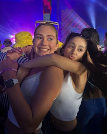 Two young adult women smile and hug at an outdoor music festival with crowd, stage lights, and yellow hat in the background.