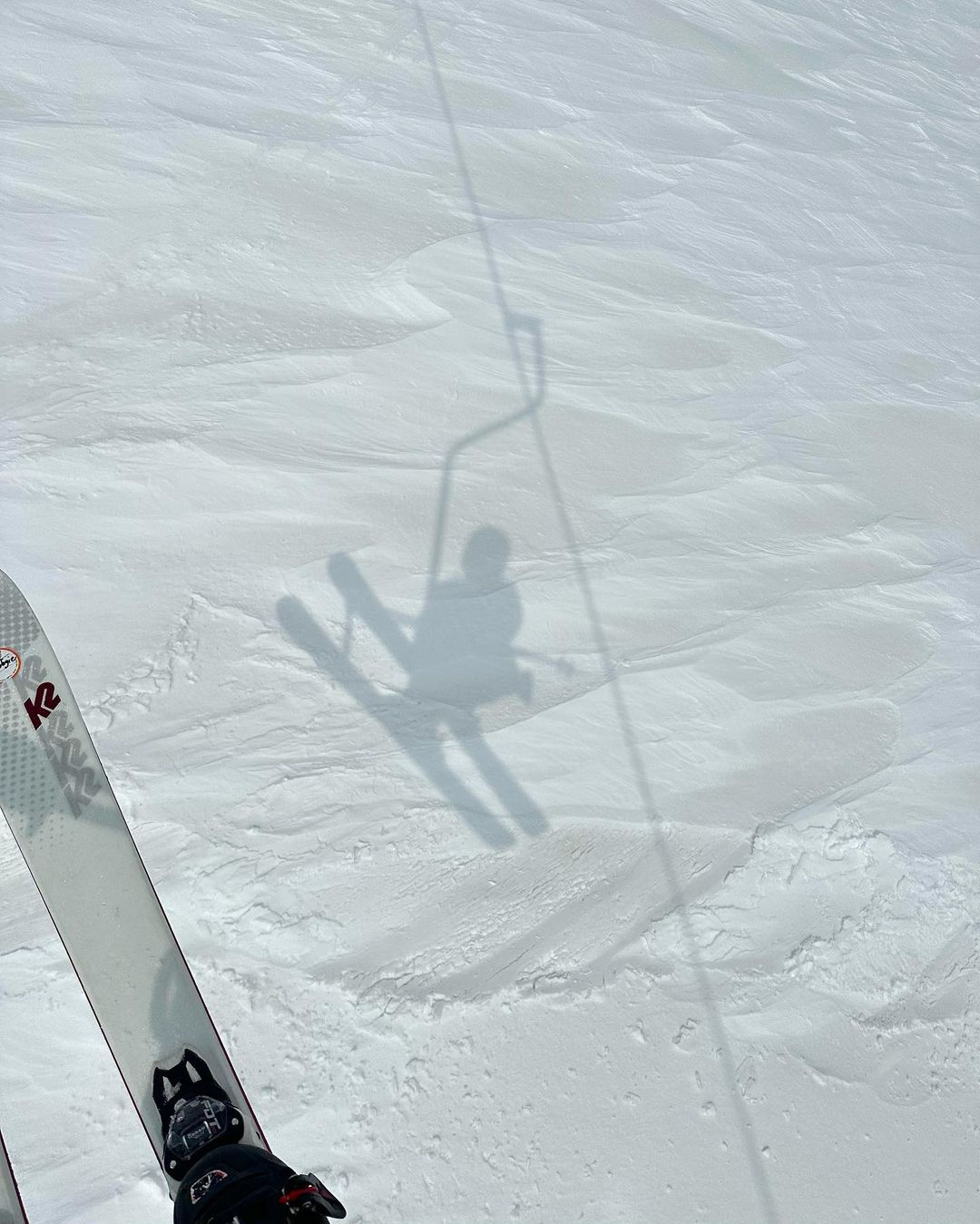 A person's shadow on skis is visible on a snowy mountain slope, with the ski lift shadow also visible in the background on a bright day.