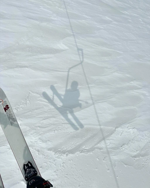 A person's shadow on skis is visible on a snowy mountain slope, with the ski lift shadow also visible in the background on a bright day.