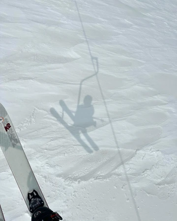 A person's shadow on skis is visible on a snowy mountain slope, with the ski lift shadow also visible in the background on a bright day.