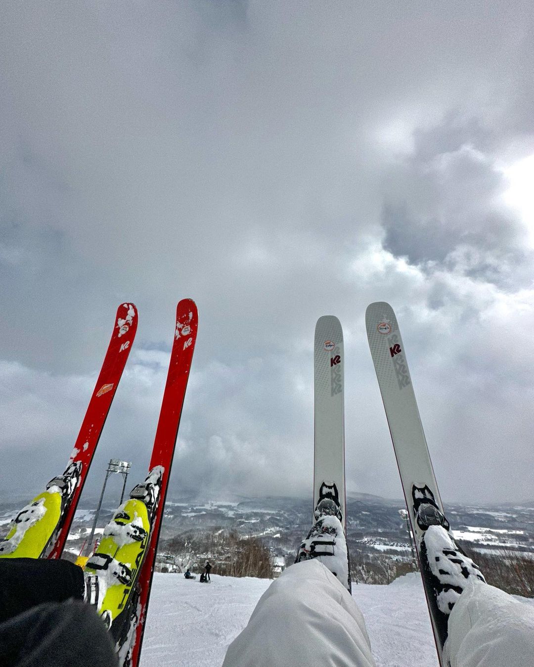 Two pairs of skis and boots rest in the snow on a mountain under a cloudy sky, suggesting a pause during a ski trip or day on the slopes.
