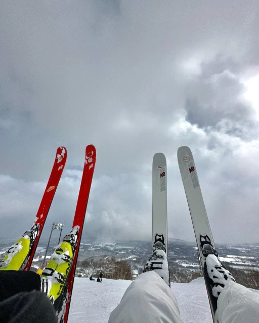 Two pairs of skis and boots rest in the snow on a mountain under a cloudy sky, suggesting a pause during a ski trip or day on the slopes.