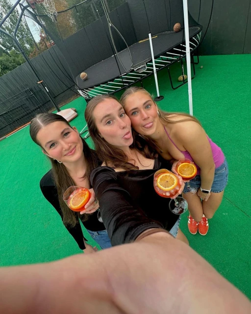 Three young women pose for a selfie, holding drinks with orange slices, outdoors with a trampoline and black fencing in the background.