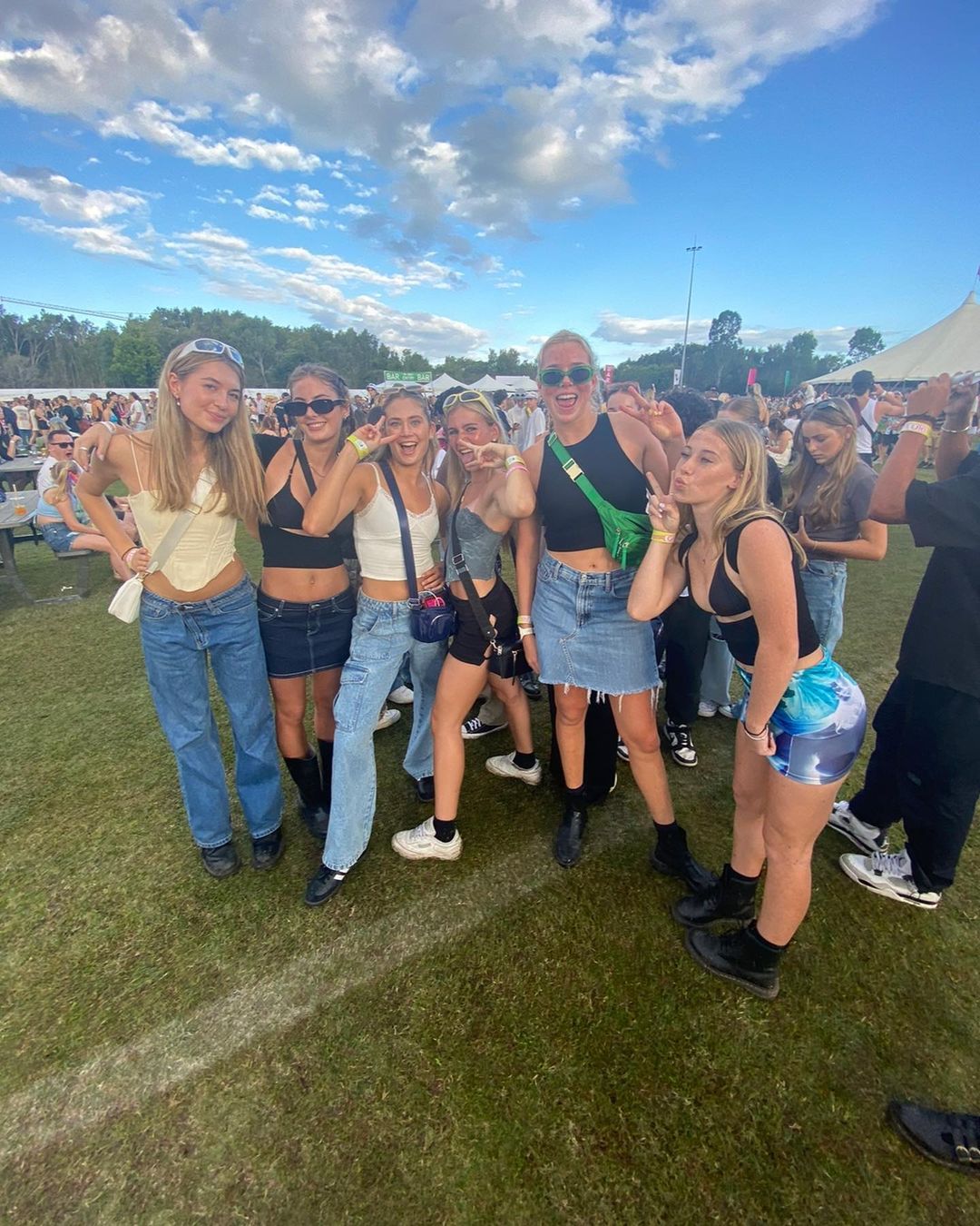 A group of six young women pose in casual summer outfits at an outdoor music festival under a sunny, partly cloudy sky.