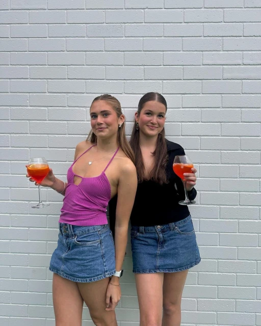 Two young women pose against a white brick wall holding vibrant orange drinks, wearing denim skirts and fashionable tops, outdoors.