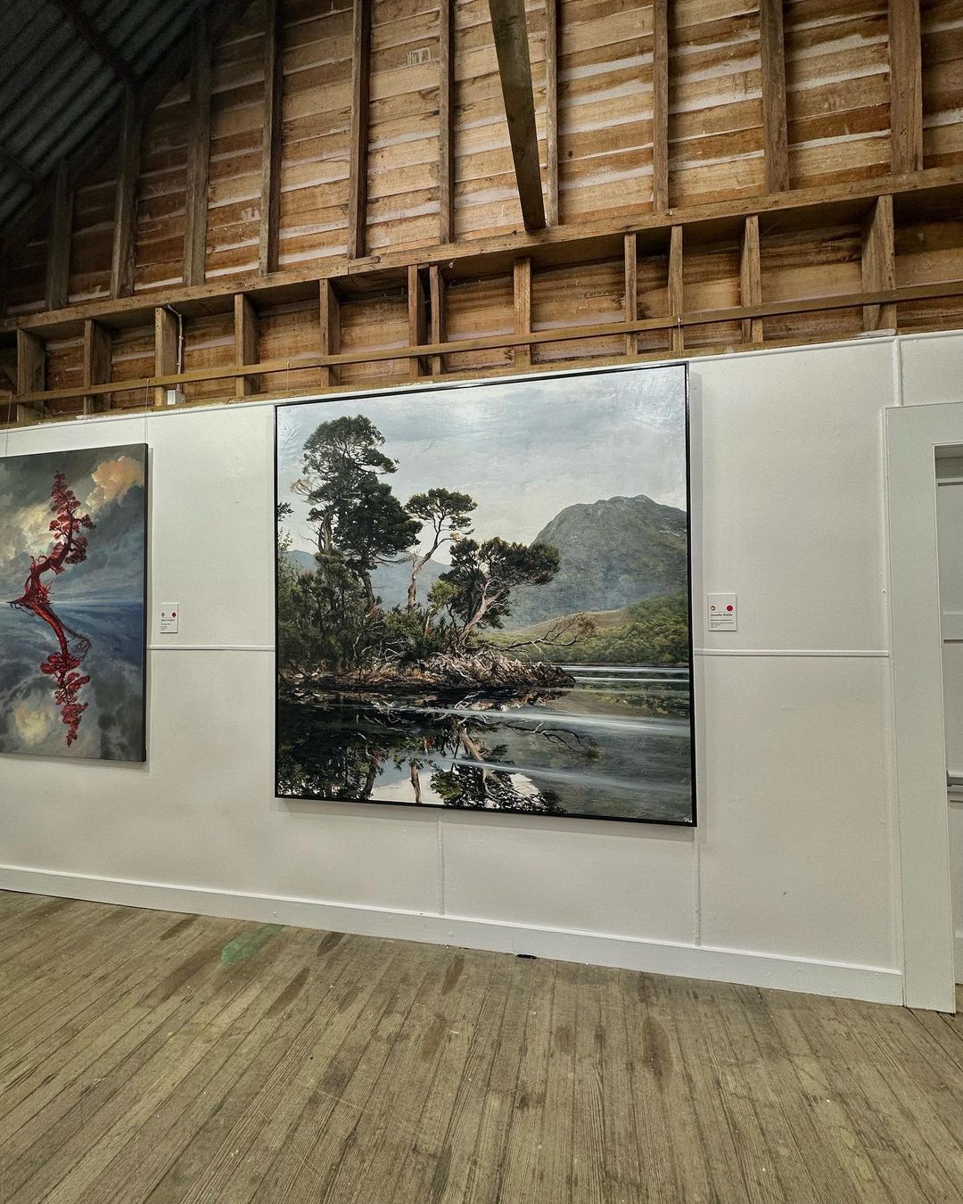 Framed painting on a white wall in a wood-beamed gallery features a landscape with trees, a mountain, and a lake reflection.