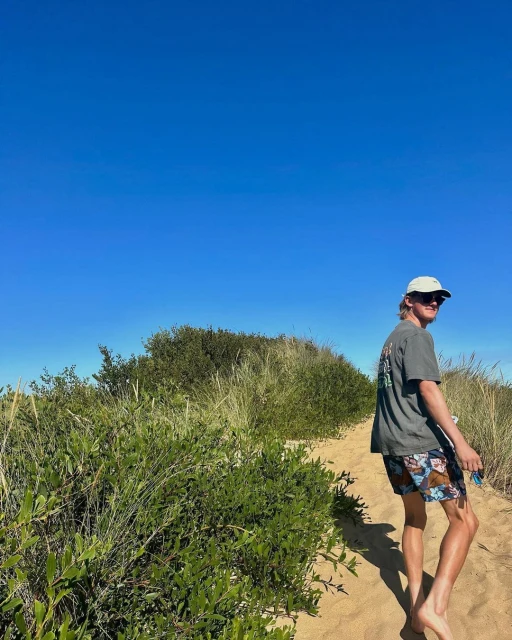 An adult male stands on a sandy path near green vegetation under a clear blue sky, wearing a cap, sunglasses, a t-shirt and shorts.