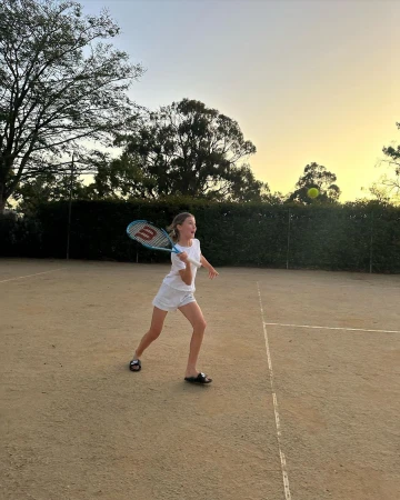 Young girl playing tennis on a clay court holding a blue racket with a yellow ball in flight above