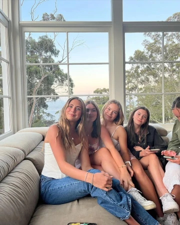 A group of five young women are sitting closely together on a couch indoors in front of a large window with a view of trees.