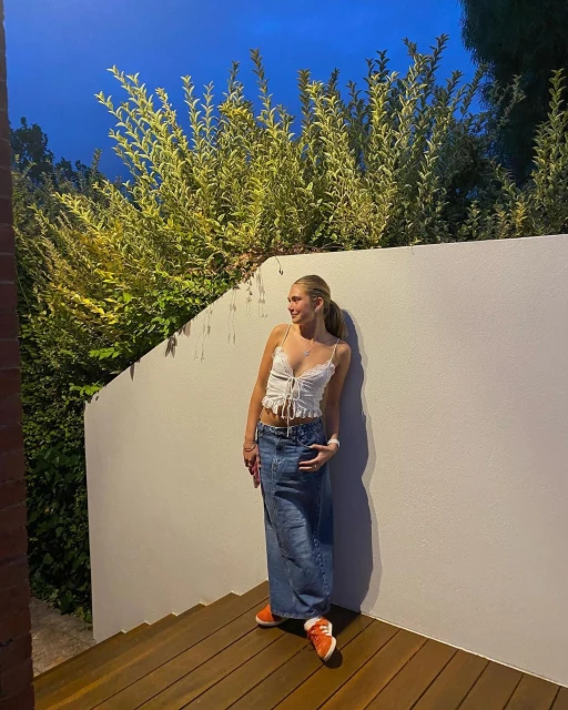 Blonde woman in white top and long denim skirt poses by a wall with green foliage in the background, wearing orange sneakers.