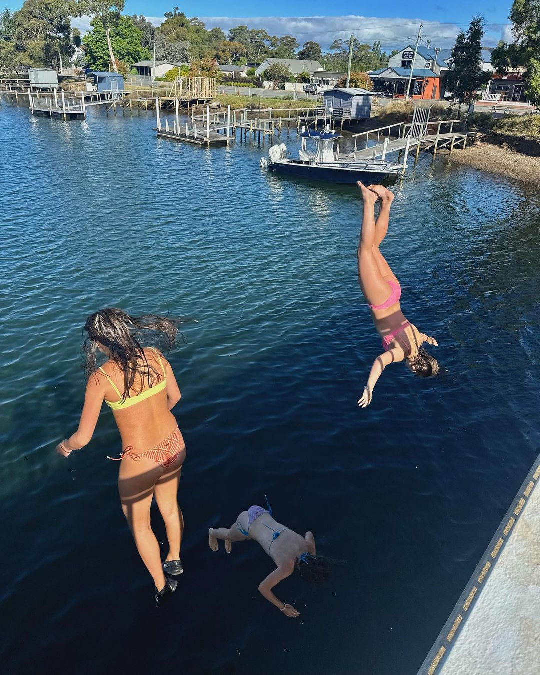 Three young women jump into the water from a dock on a sunny day, with boats, houses, and trees in the background under a blue sky.
